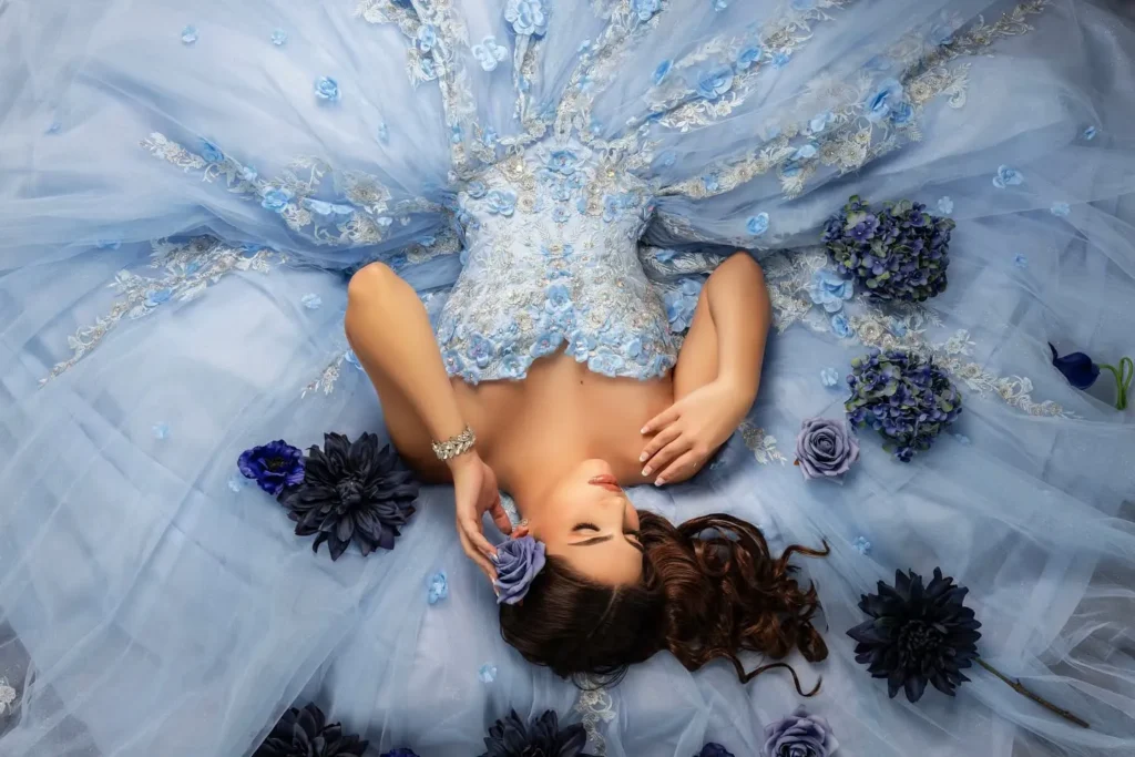Quinceañera con vestido azul acostada entre flores en sesión artística