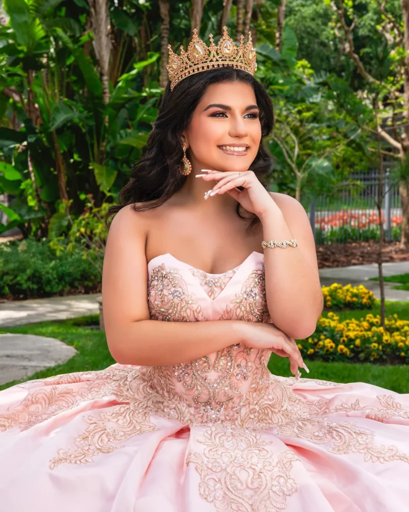Joven con vestido de quince años rosado y corona dorada posando sonriente en jardín tropical